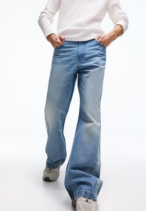 Osoba oblečená v svetlomodrých džínsach so širokými nohavicami, bielom dlhorukávovom tričku a bielych teniskách, stojaca s rukami v kapse, na bielom pozadí.