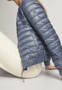 Personne assise par terre portant une doudoune matelassée bleue et un pantalon écru, seuls le torse et les jambes sont visibles.