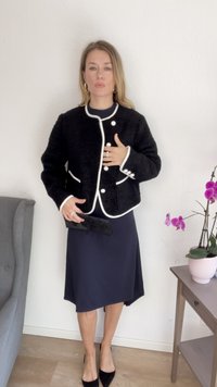 Μαύρο cropped σακάκι με λευκές λεπτομέρειες και κουμπιά, φορεμένο πάνω από ένα ναυτικό φόρεμα. Το μοντέλο κρατάει μια μαύρη τσάντα clutch, ενώ στέκεται δίπλα σε μια ορχιδέα σε γλάστρο.