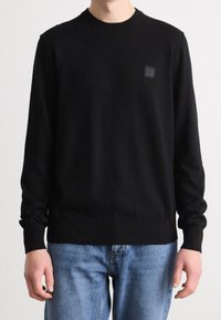 Personne portant un pull noir uni à manches longues avec un petit logo carré sur la poitrine et un jean bleu clair, debout devant un fond blanc.