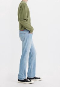 Vaqueros de mezclilla azul claro con corte recto, combinados con una camisa de manga larga suelta de color verde oliva y zapatillas negras con suela blanca.