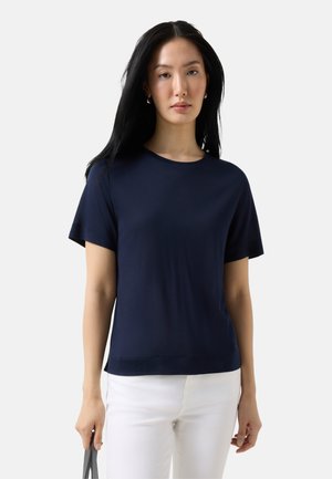 Vrouw met zwart lang haar, gekleed in een marineblauw T-shirt met korte mouwen en een witte broek, die met haar linkerhand een grijze tas vastheeft.