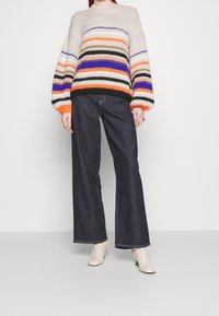 Pull en tricot avec des rayures horizontales multicolores en orange, violet et vert, associé à un jean large en denim foncé et des bottines beiges.
