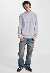 Grå hættejakke med lynlås og teksten "SPORTSWEAR", kombineret med falmede blå jeans og sorte sneakers. Indeholder teksturerede ærmer og en afslappet pasform.