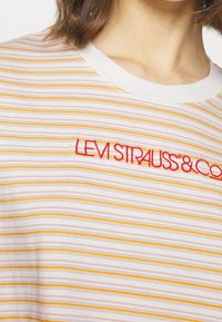 Maglietta bianca con strisce orizzontali gialle e arancioni, con logo ricamato rosso "LEVI STRAUSS & CO" vicino allo scollo.