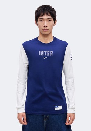 Ung mand iført en blå og hvid langærmet skjorte med "INTER" og et Nike-logo på brystet, stående mod en ensfarvet baggrund.