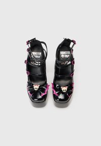 Pantofi cu toc platform din piele lacuită neagră, cu multiple curele, o aplicație în formă de fundiță reprezentând un craniu și aripi de lilieci în nuanțe de roz și mov.