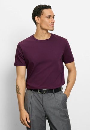 Mann mit kurzen lockigen Haaren, der ein violettes kurzärmeliges Hemd trägt, das in graue Hosen mit schwarzem Gürtel gesteckt ist, steht mit einer Hand in der Tasche.