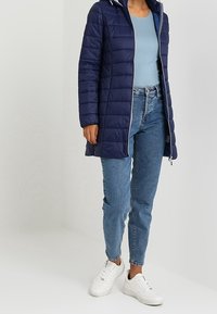 Abrigo acolchado azul marino con capucha, cierre de cremallera y textura acolchada, combinado con una camiseta ajustada azul claro y jeans de mezclilla, completado con zapatillas blancas.