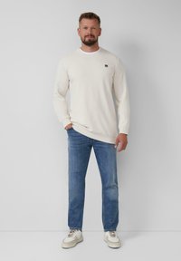 Sweatshirt beige clair à col ras du cou avec des poignets côtelés, associé à un jean droit bleu et des baskets blanches. Détail de logo discret sur la poitrine.