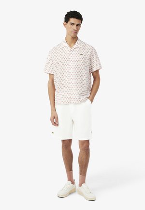 Giovane uomo con una camicia a maniche corte con bottoni e fantasia rosa, pantaloncini bianchi, calzini rosa e scarpe da ginnastica bianche, in piedi con una mano in tasca.