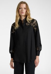 Camicia nera con bottoni, con dettagli in pizzo sulle maniche. Presenta un design strutturato e un tessuto texture, abbinata a denim scuro.