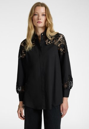 Camicia nera con bottoni, con dettagli in pizzo sulle maniche. Presenta un design strutturato e un tessuto texture, abbinata a denim scuro.