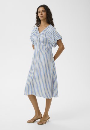 Femme debout portant une robe mi-longue à rayures verticales bleues, jaunes et blanches avec des manches courtes bouffantes, sur un fond uni.