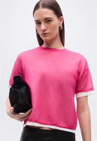 Γυναίκα με ίσια καστανά μαλλιά φοράει ασημένια σκουλαρίκια κρίκους και ένα ροζ διχτυωτό κοντομάνικο τοπ, κρατώντας μια μικρή μαύρη τσάντα μπροστά σε λευκό φόντο.