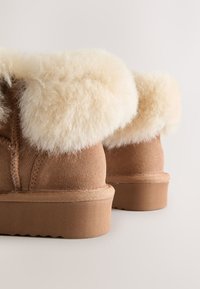 Stivaletti in pelle scamosciata color marrone chiaro con polsino in shearling morbido di colore crema. Suola piatta in gomma con texture e dettagli di cuciture visibili.