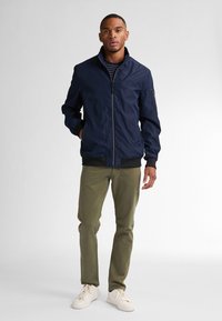 Marineblauwe bomberjack met een hoge kraag, frontale rits en geribde manchetten. Draagt over olijfgroene broek en witte sneakers.