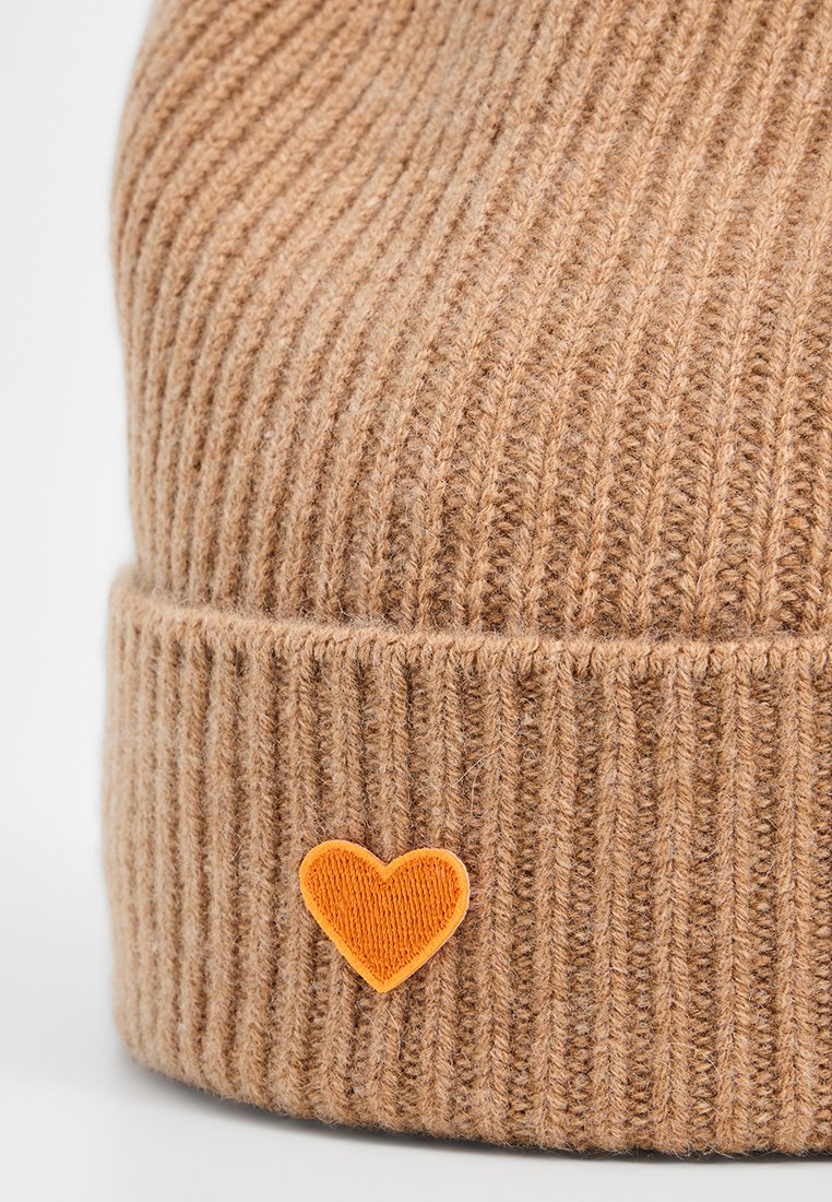 Jasnobrązowa, przędzy rypsowej czapka z małą, pomarańczową łatą w kształcie serca na złożonym mankiecie.