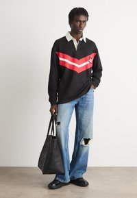 Una persona in piedi indossa una maglietta nera a maniche lunghe con strisce rosse, jeans blu strappati, scarpe nere, e tiene una grande borsa nera contro un muro bianco.