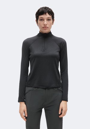 Jong persoon met kort donker haar, gekleed in een zwart langarmig topje met rits en een donkere broek, staand tegen een effen achtergrond.