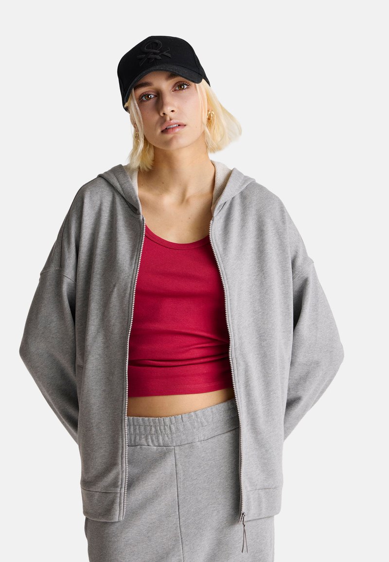 Felpa grigia con zip e texture morbida, indossata sopra una canotta rossa. Accessoriata con un cappellino nero con logo. Vestibilità casual e rilassata.