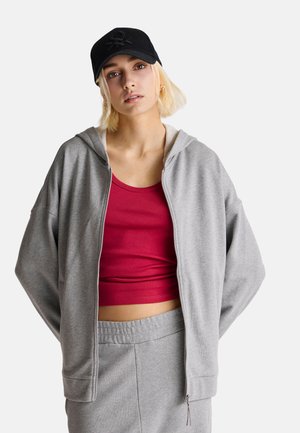 Grijze zip-up hoodie met een zachte textuur, gedragen over een rode tanktop. Gecombineerd met een zwarte pet met een logo. Casual, relaxed fit.