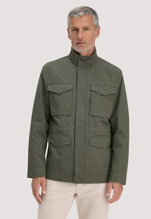 Mann mit grauem Haar und Bart, der eine olivgrüne Jacke mit vier vorderen Klappentaschen und beigefarbene Hose trägt, steht vor einem schlichten Hintergrund.