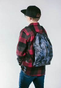 Zaino con cordoncino di colore nero con un motivo testurizzato, indossato da una persona che porta una camicia a quadri rossa e nera e jeans.