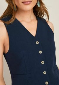 Vrouw in een mouwloze marineblauwe jurk met een V-hals, vijf knopen aan de voorkant en kleine zakdetails.