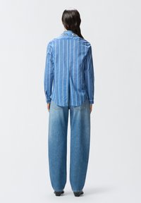 Camicia blu a righe con bottoni e spacco sul retro, abbinata a jeans in denim blu chiaro a gamba larga. Design semplice con vestibilità casual e texture morbida.