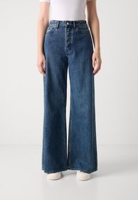 Un pantalon en denim bleu, taille haute et pattes d'éléphant. Il comporte des poches avant, une fermeture à bouton et un ourlet effiloché. Associé à des baskets blanches.