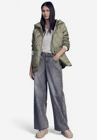 Giacca trapuntata verde con cappuccio, maglietta bianca a maniche lunghe e jeans larghi grigi. Le scarpe platform beige chiaro completano l'outfit.