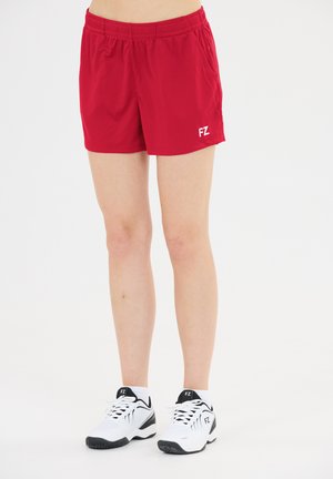 Person iført røde sportsshorts og hvide sneakers med sorte detaljer, stående mod en ensfarvet hvid baggrund.