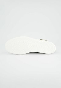 Semelle de sneaker blanche avec un motif texturé, arborant un logo bien en relief verticalement sur le côté. Le matériau semble en caoutchouc.