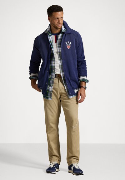Mand iført et lagdelt outfit med en marineblå jakke mærket USA, grønternet skjorte, hvid T-shirt, beige bukser og marineblå sneakers, stående mod en ensfarvet baggrund.
