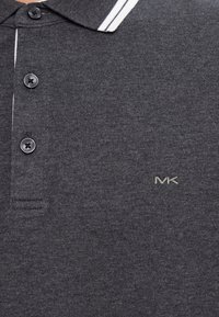 Tmavosivá polo košeľa s čiernymi gombíkmi, bielym prúžkom okolo goliera a malým vyšitým logom "MK" na hrudi.