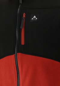 Svart och röd fleecejacka med dragkedja, som har en bröstficka och logotyp. Texturerad kropp och släta ärmar.