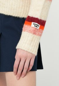 Sweter dzianinowy w kremowym kolorze z wielokolorowymi paskami na rękawach (czerwony, pomarańczowy, różowy) i ściągaczami; ma metkę "COLOURFUL REBEL".