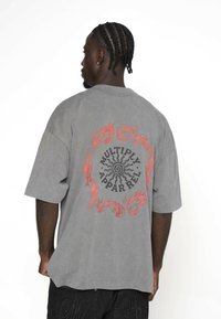 Szara, oversize'owa koszulka z okrągłym czarnym i czerwonym wzorem graficznym na plecach, z płomieniami wokół napisu "MULTIPLY APPAREL."