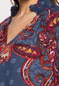 Blusa azul marino con intrincados patrones de paisley en rojo y amarillo, cuello con volantes y tejido texturizado. Presenta detalles florales y acentos en contraste.