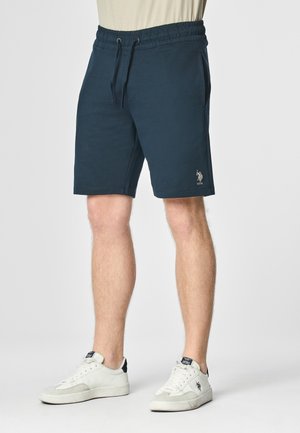 Man draagt marineblauwe US Polo Assn. shorts met elastische tailleband en witte sneakers met zwarte logo-accenten.
