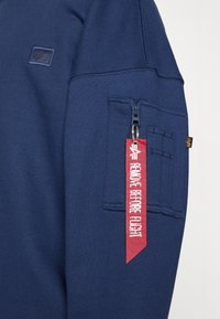 Felpa blu navy con tasca con zip, caratterizzata da un'etichetta rossa con la scritta "RIMUOVERE PRIMA DEL VOLO" e dettagli in tessuto testurizzato.