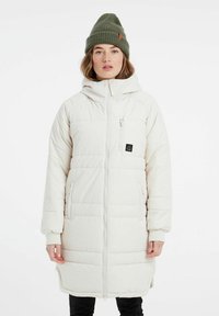 Bílý prošívaný kabát s kapucí, má zapínání na zip, boční kapsy a kapsu na hrudi. Doplněný o zelenou pletenou čepici.
