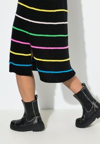 Schwarzes Strickkleid mit mehrfarbigen horizontalen Streifen. Das Model trägt schwarze Knöchelschuhe mit Nietenakzenten und einer dicken Sohle.