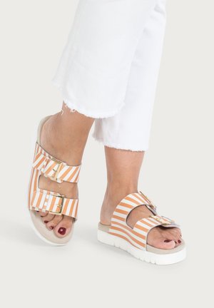 Füße, die orange-weiß gestreifte Sandalen mit goldenen Schnallen und weißen Plateausohlen tragen, kombiniert mit weißen kurzen Hosen.