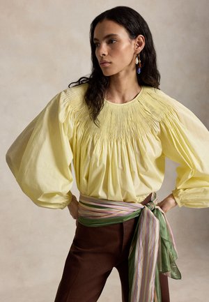 Naisella on tummat aaltoilevat hiukset, hänellä on päällään keltainen kerrostettu paita, jossa on liehukkeet hihnoissa, ruskeat housut ja raidallinen vihreä- ja vaaleanpunainen vyötäröhuivi, joka on sidottu rusetille.