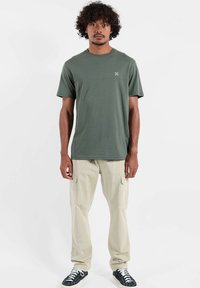 T-shirt vert olive à manches courtes avec col rond, associé à un pantalon cargo beige avec poches latérales et des baskets noires.