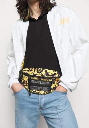 Osoba ubrana w białą kurtkę na czarnej koszulce, z czarno-złotą nerka Versace Jeans Couture i jasnoniebieskimi dżinsami.