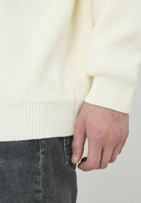 Pull en maille côtelée blanc avec une finition texturée, arborant un petit logo sur le côté. Porté avec un jean gris foncé. Main visible.