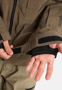 Hände, die den Klettverschluss des Ärmels einer braunen GORE-TEX-Jacke anpassen, wobei das Daumenloch und ein Ring am linken Mittelfinger zu sehen sind.